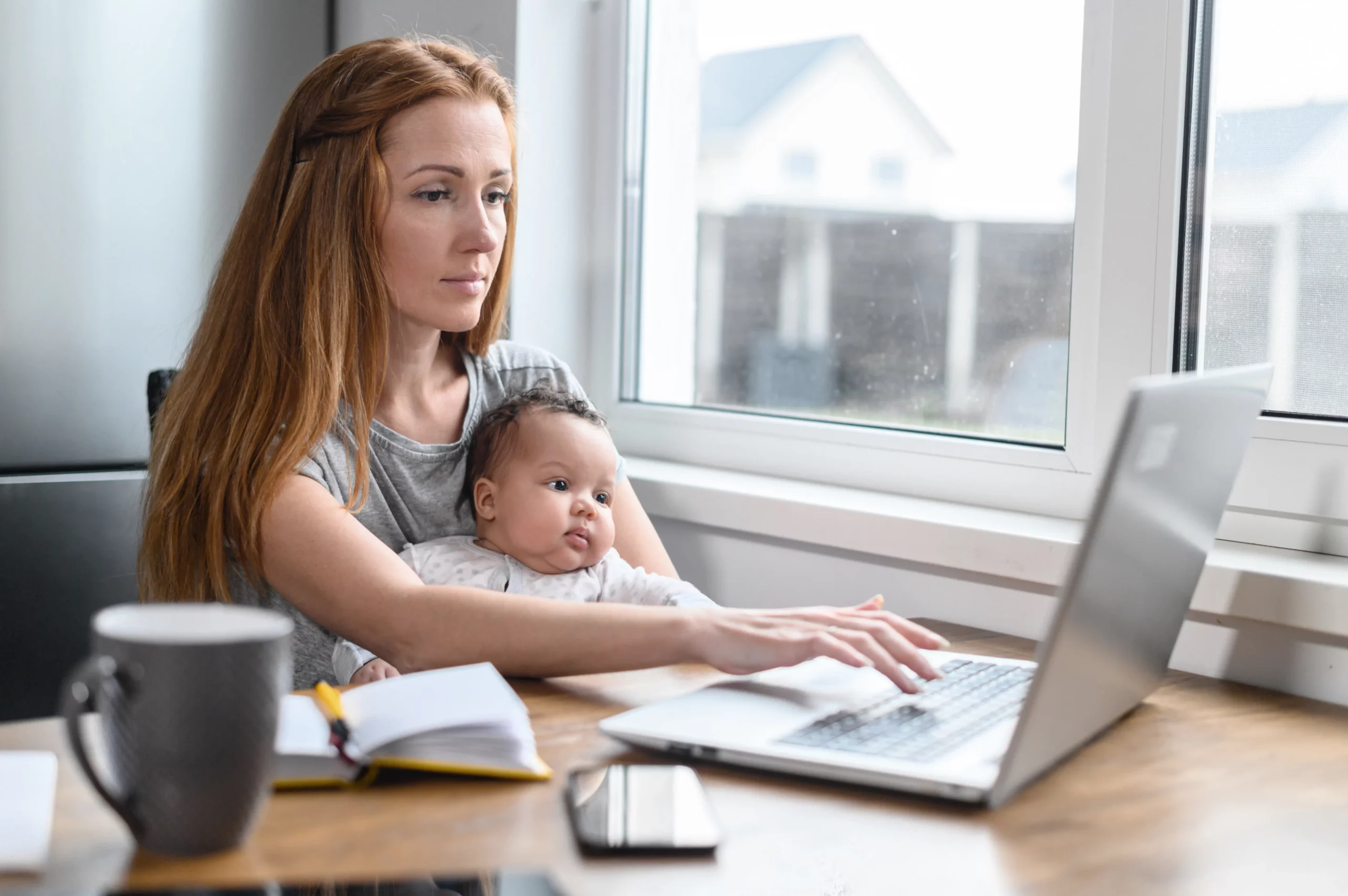 Woman working on laptop while holding baby on her lap looking miserable