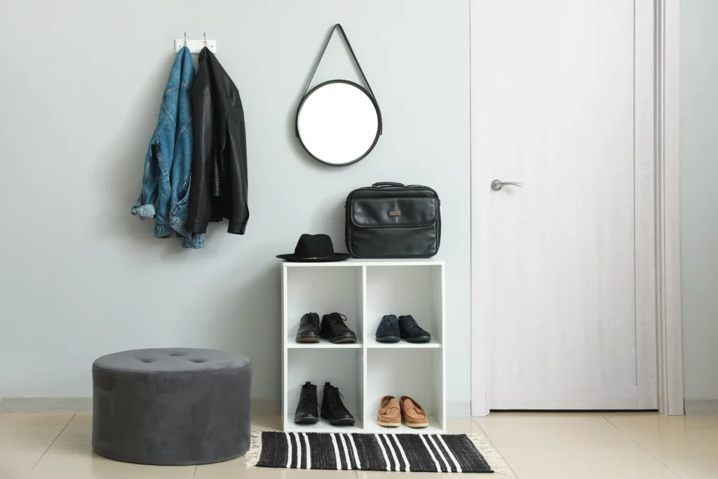 Shoe rack at entry way, under circular mirror next to ottoman, and near hooks with jackets on them